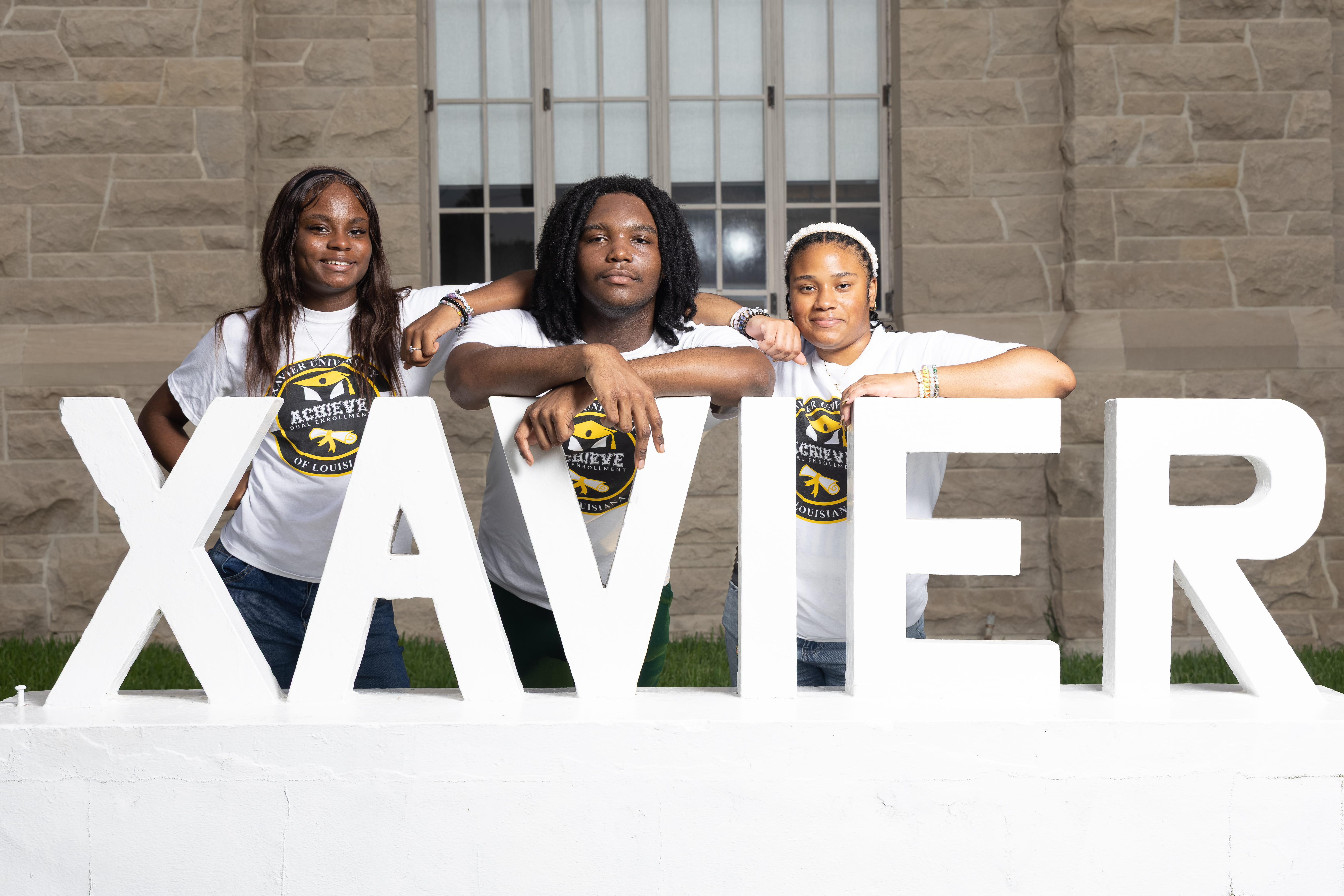 Three Achieve Dual Enrollment students, two Black female students and one Black male student, pose behind the Xavier sign in front of the Xavier University of Louisiana Administration Building.